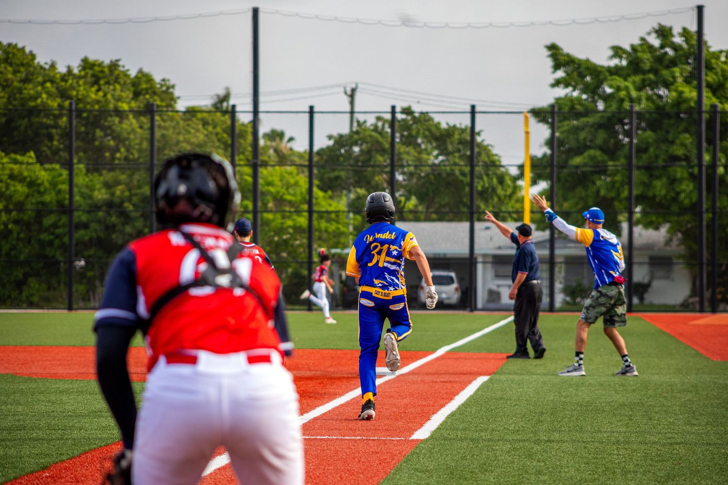 Baseball players compete on a red turf field, with a batter running bases and coaches signaling during an outdoor game.