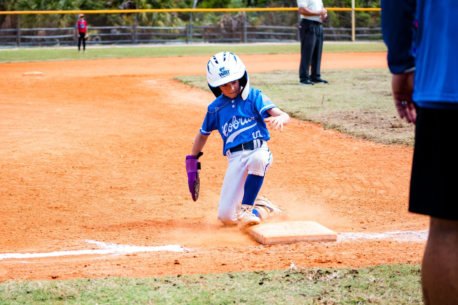 A baseball player slides into home base during a youth league game with a cloud of dust rising from the dirt.