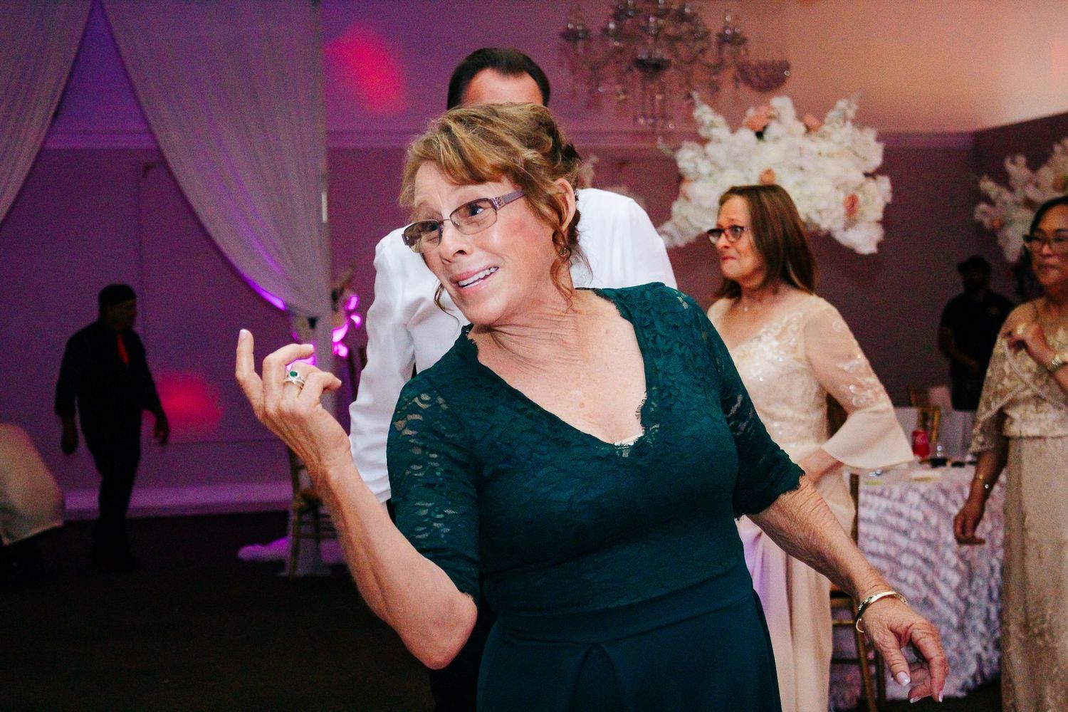 Wedding reception guests dance and celebrate on a dimly lit dance floor with purple ambient lighting.