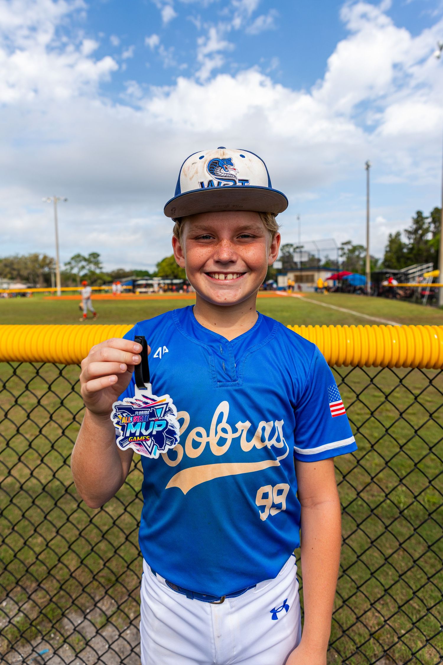 Young baseball player in blue and white uniform stands by chain link fence at sunny baseball field.