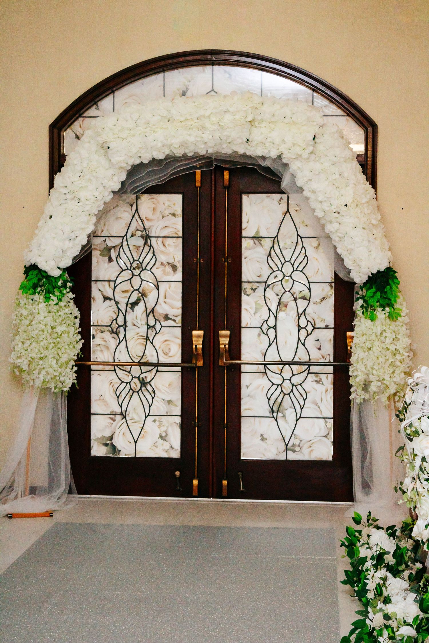 Elegant double doors with stained glass framed by a white floral arch and greenery.