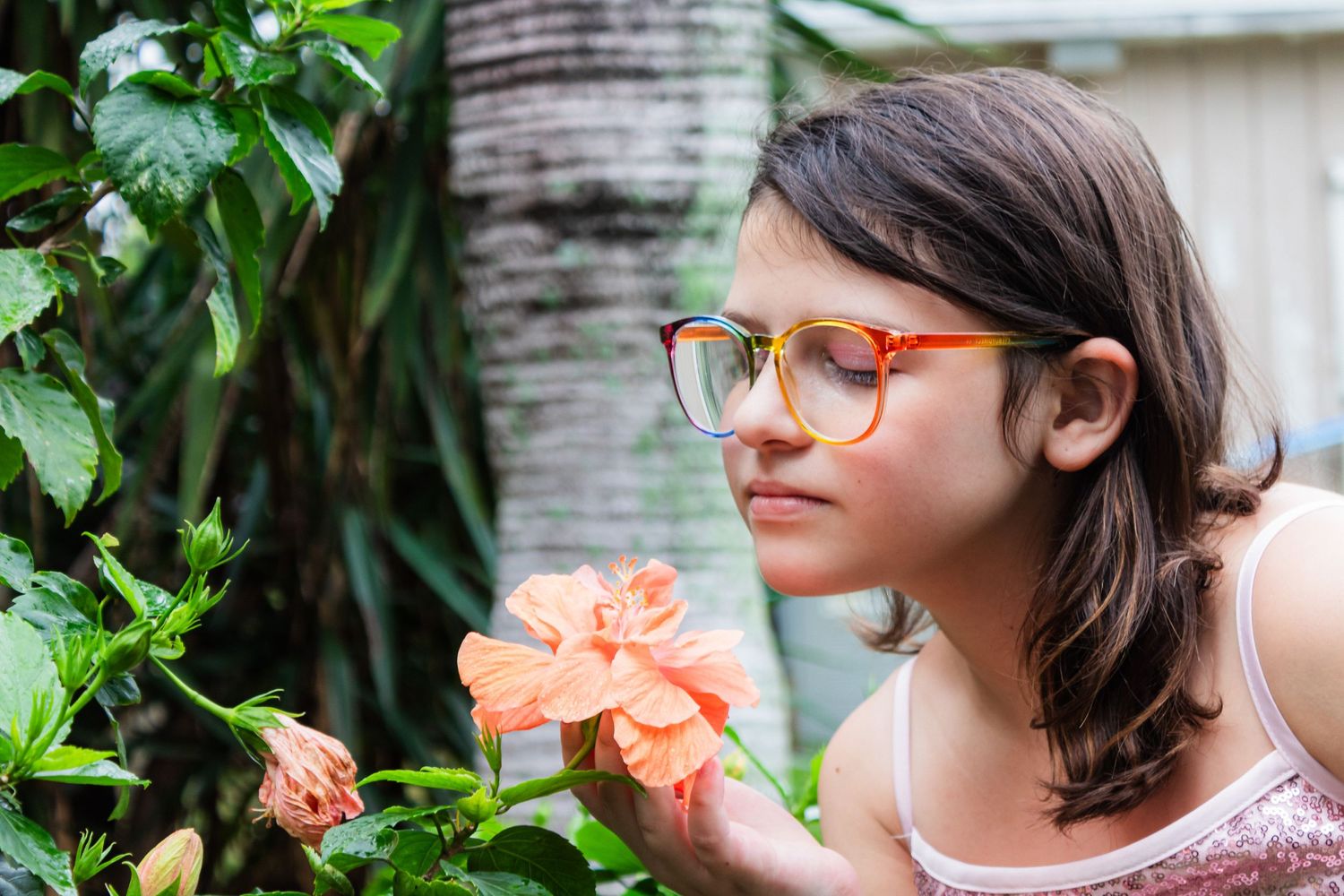 Person in glasses smelling coral-colored flower in tropical garden setting.