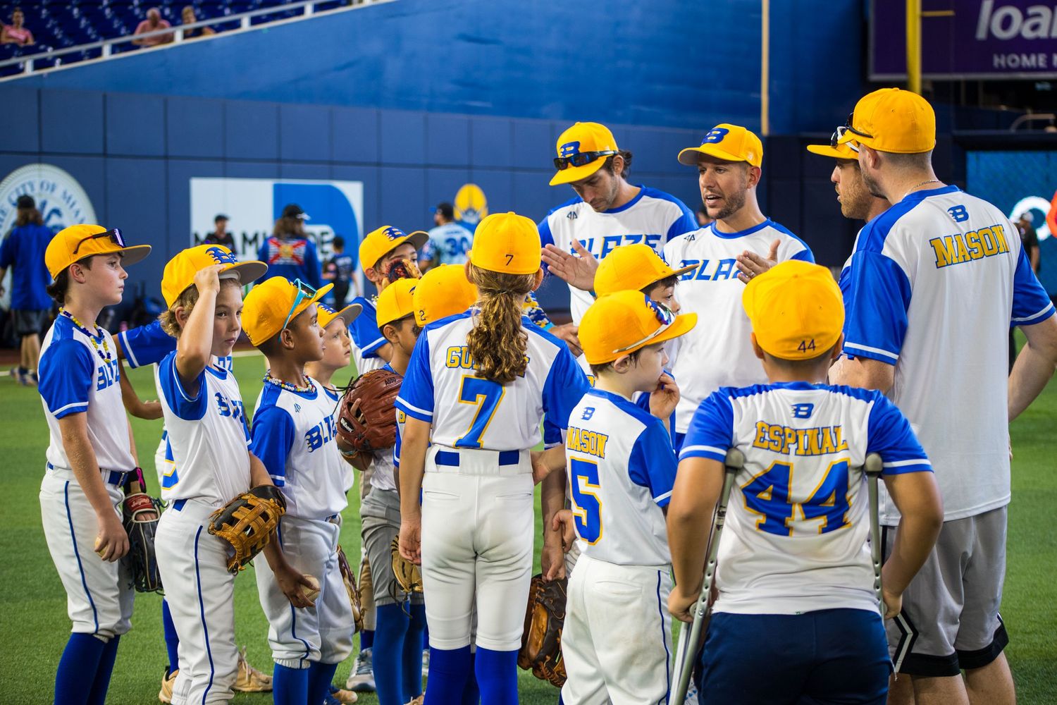 Youth baseball team in blue and white uniforms with yellow caps gather for a group huddle on the field.