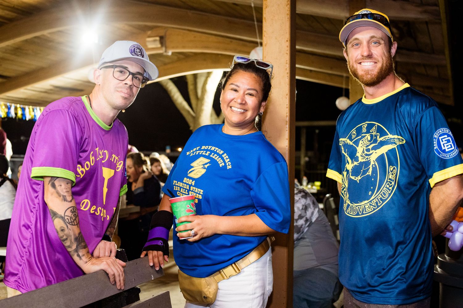 Three people wearing sports jerseys in purple, blue, and teal colors smile at a baseball event.