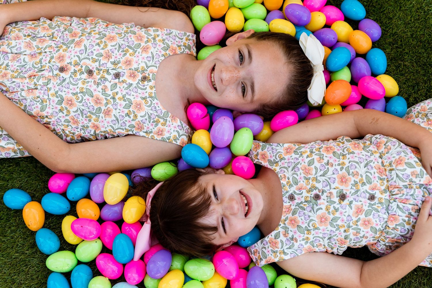 Two figures in floral dresses lying on grass surrounded by colorful plastic Easter eggs creating a vibrant display.