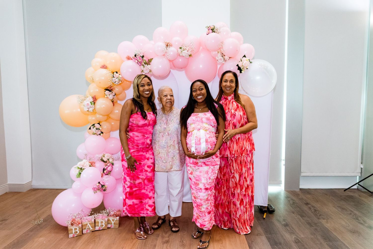 A group poses in front of a pink and peach balloon arch at a baby shower celebration.