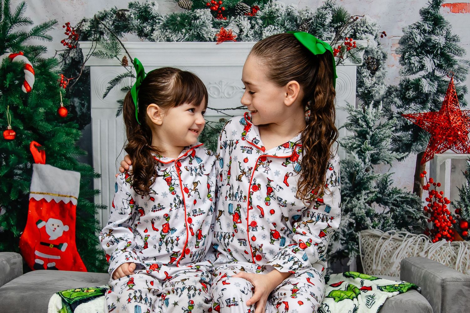 Two children in matching holiday pajamas smile at each other in front of a festive Christmas backdrop with garland and decor.