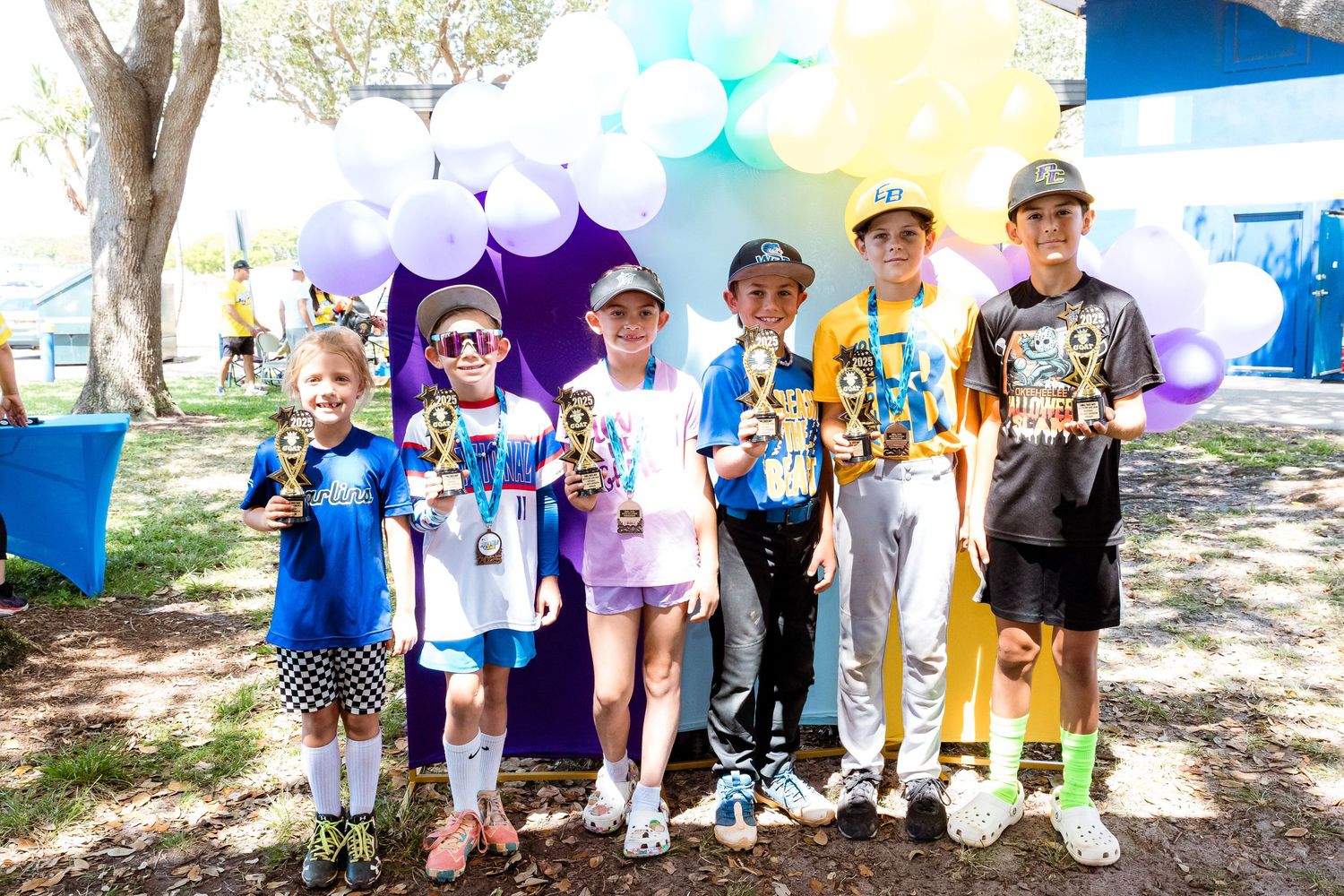 Children in casual attire stand together at an outdoor event with purple and blue balloons in the background.