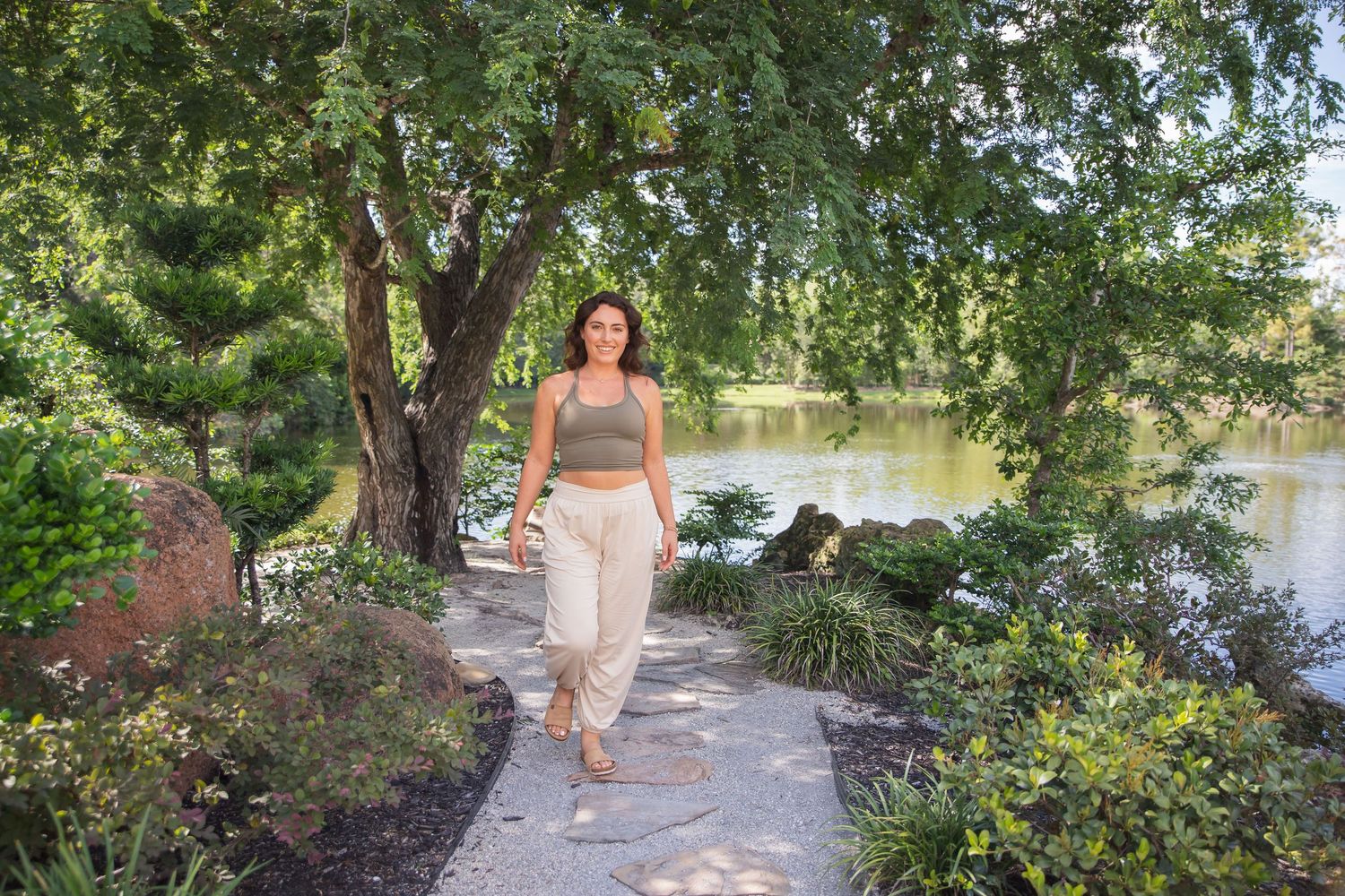 A person walks along a tree-lined path beside a lake on a sunny summer day.