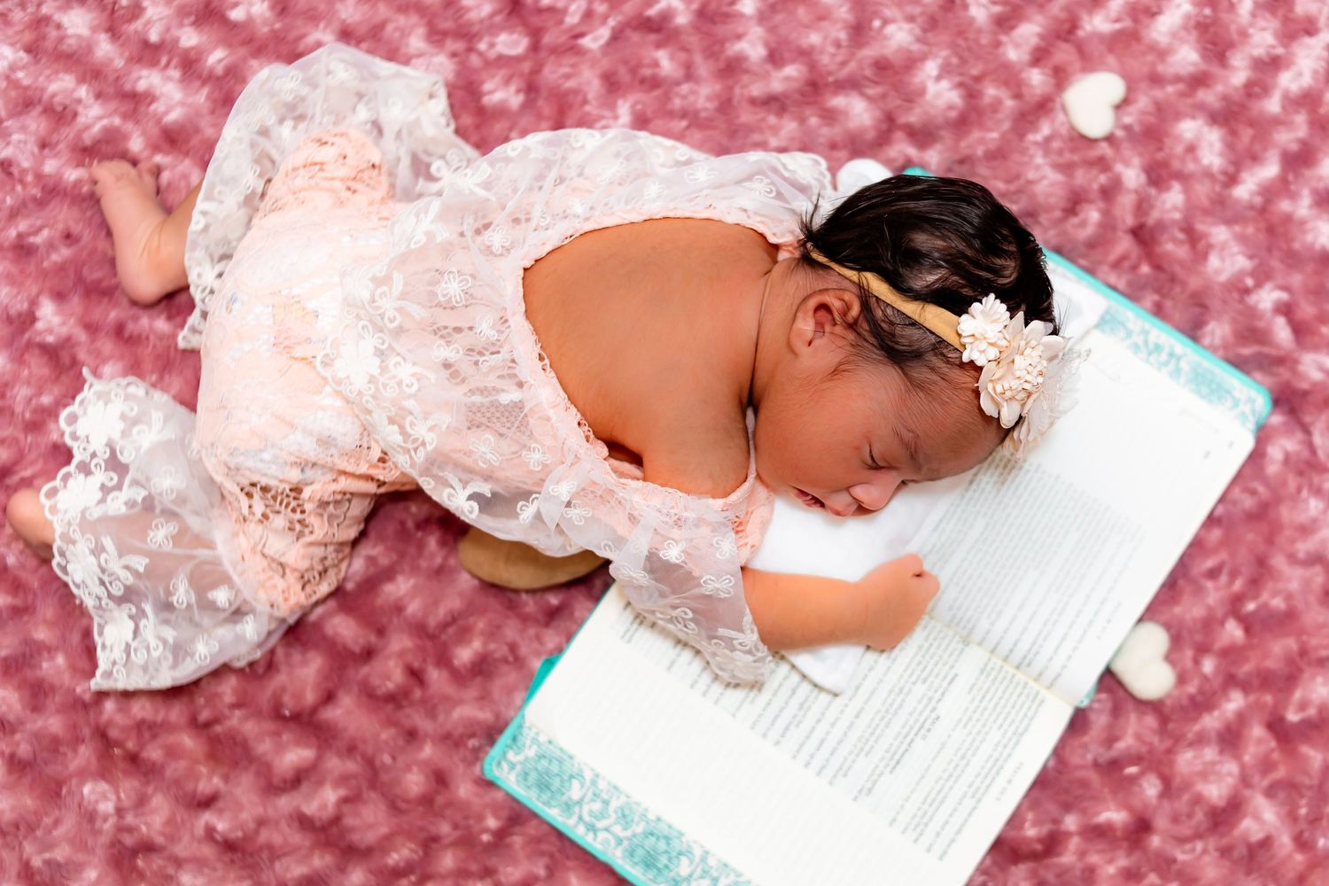Newborn baby girl in pink lace outfit with floral headband sleeping on birth certificate with rose petals.