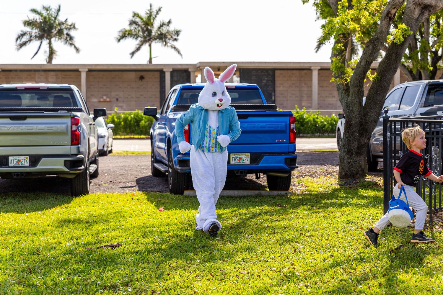 Person in Easter Bunny costume stands on green lawn near parked trucks while a child runs nearby on a sunny day.