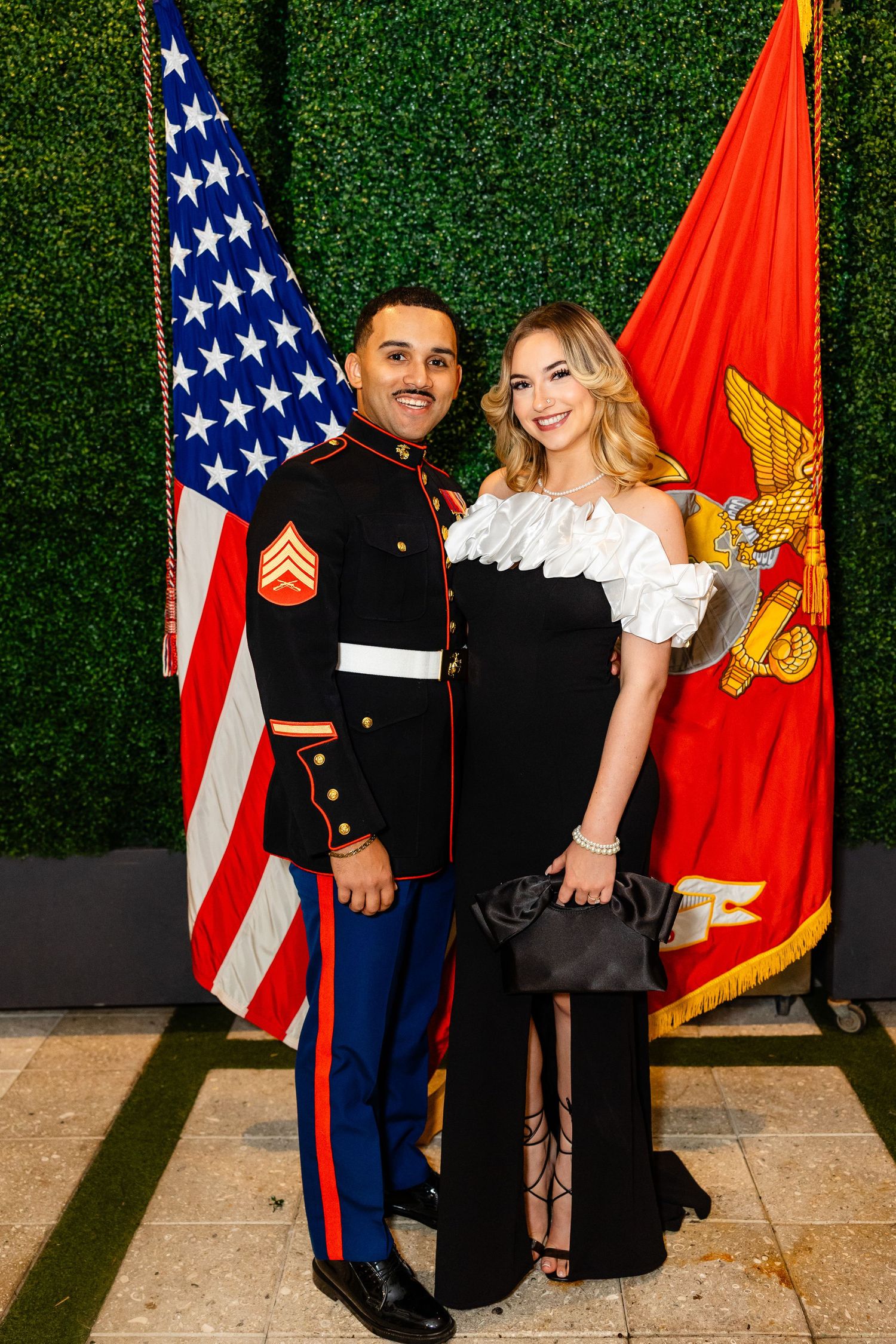 A couple stands together at a formal event with American and Marine Corps flags against a green hedge wall.