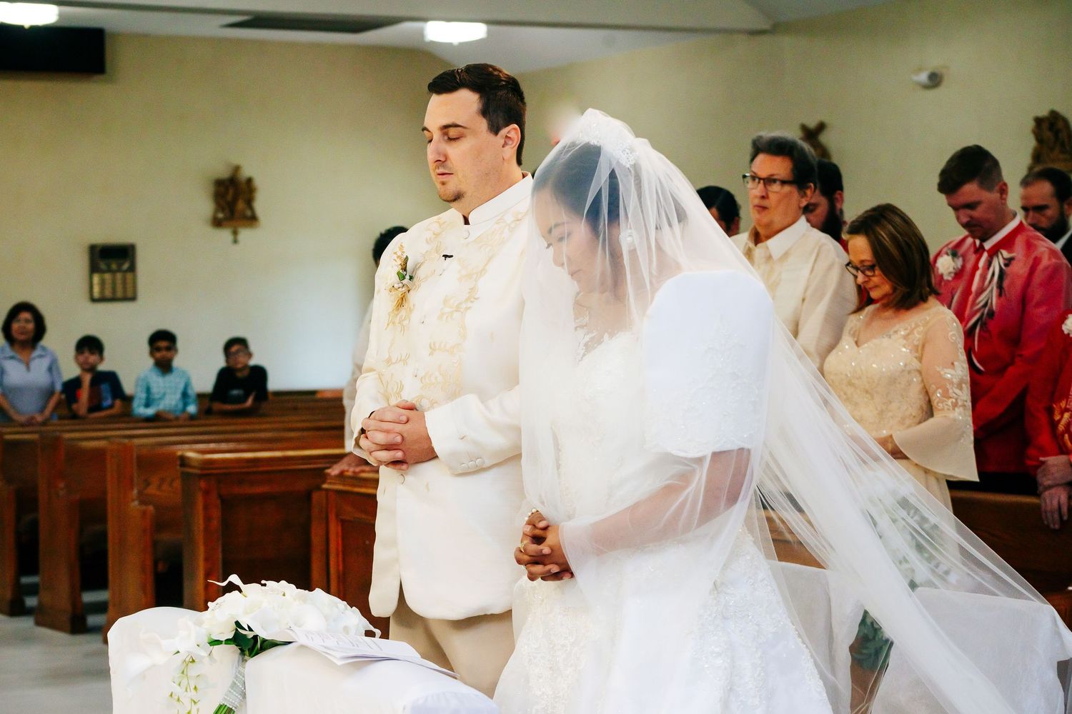 A couple holds hands during their Catholic church wedding ceremony as guests watch from wooden pews.