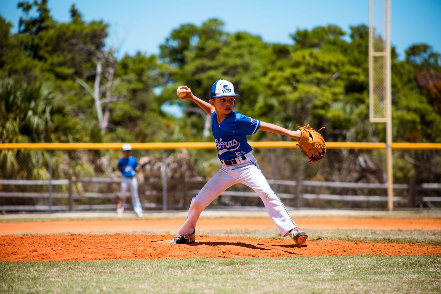 Youth baseball pitcher in a blue jersey winding up to throw from the mound in a sunny stadium.