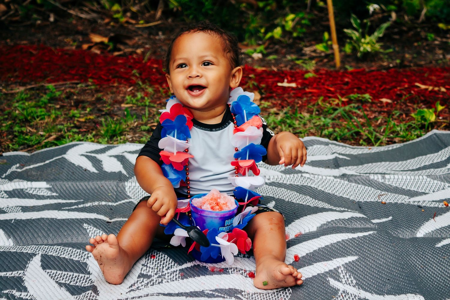 Baby wearing blue and red lei necklace sits smiling on patterned blanket outdoors.