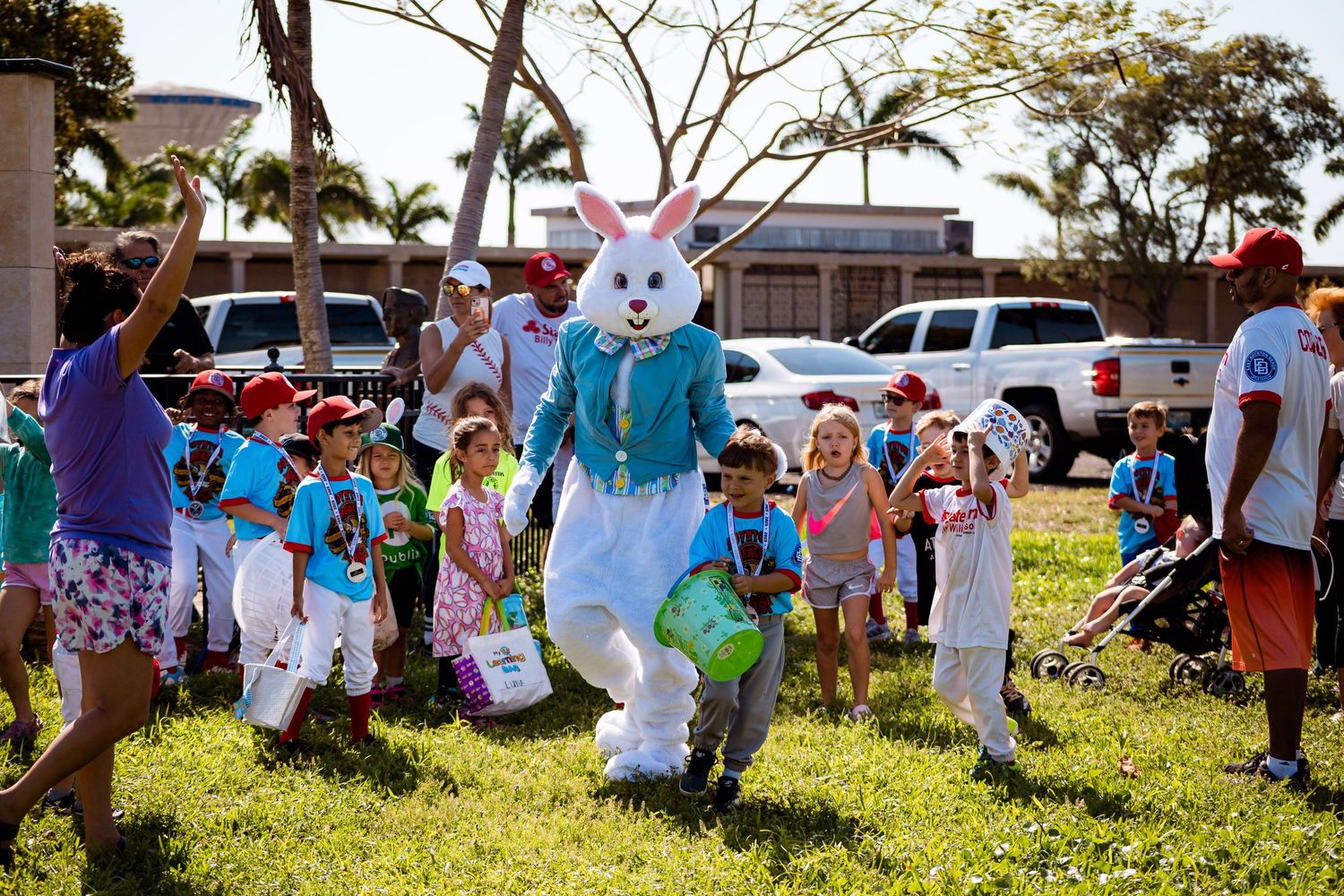 Children run and play excitedly with the Easter Bunny mascot on a sunny grass lawn during a holiday celebration.