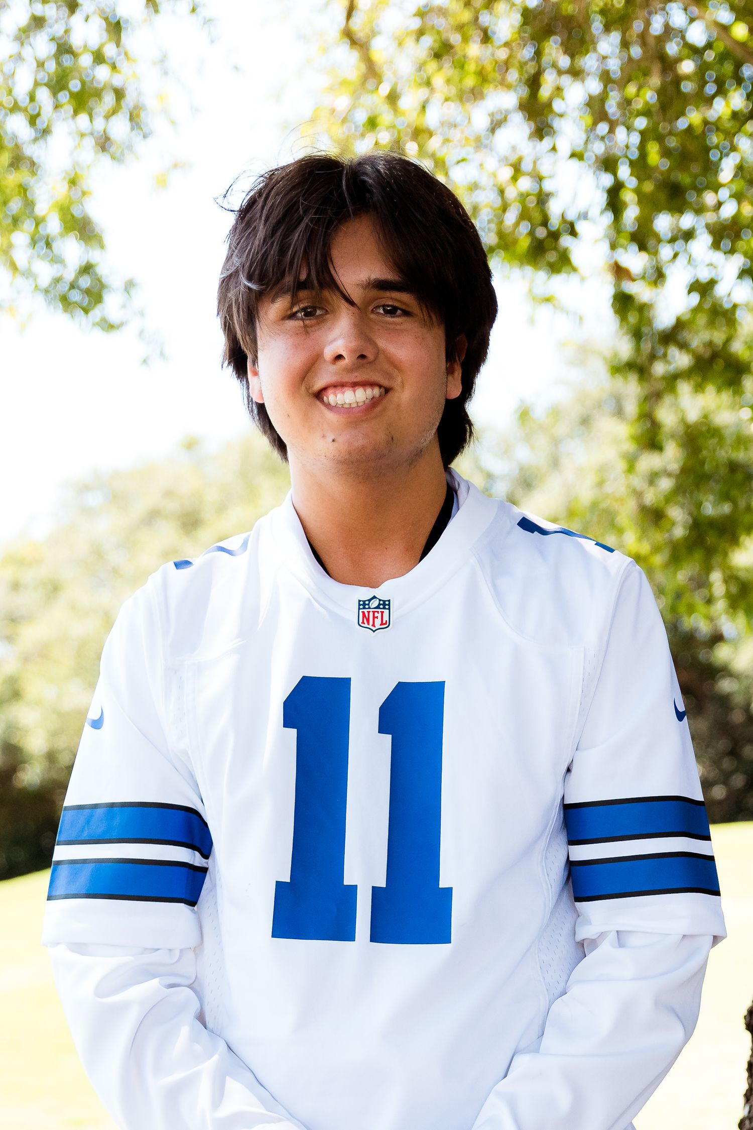 A person wearing a white Dallas Cowboys NFL jersey number 11 poses outdoors with trees in the background.