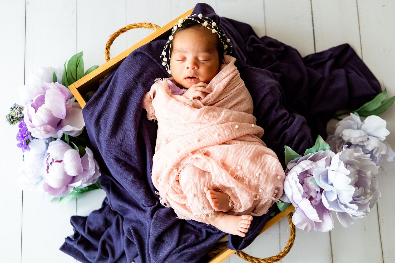 Newborn baby swaddled in pink wrap sleeping in a basket with purple floral decor and navy blue fabric.