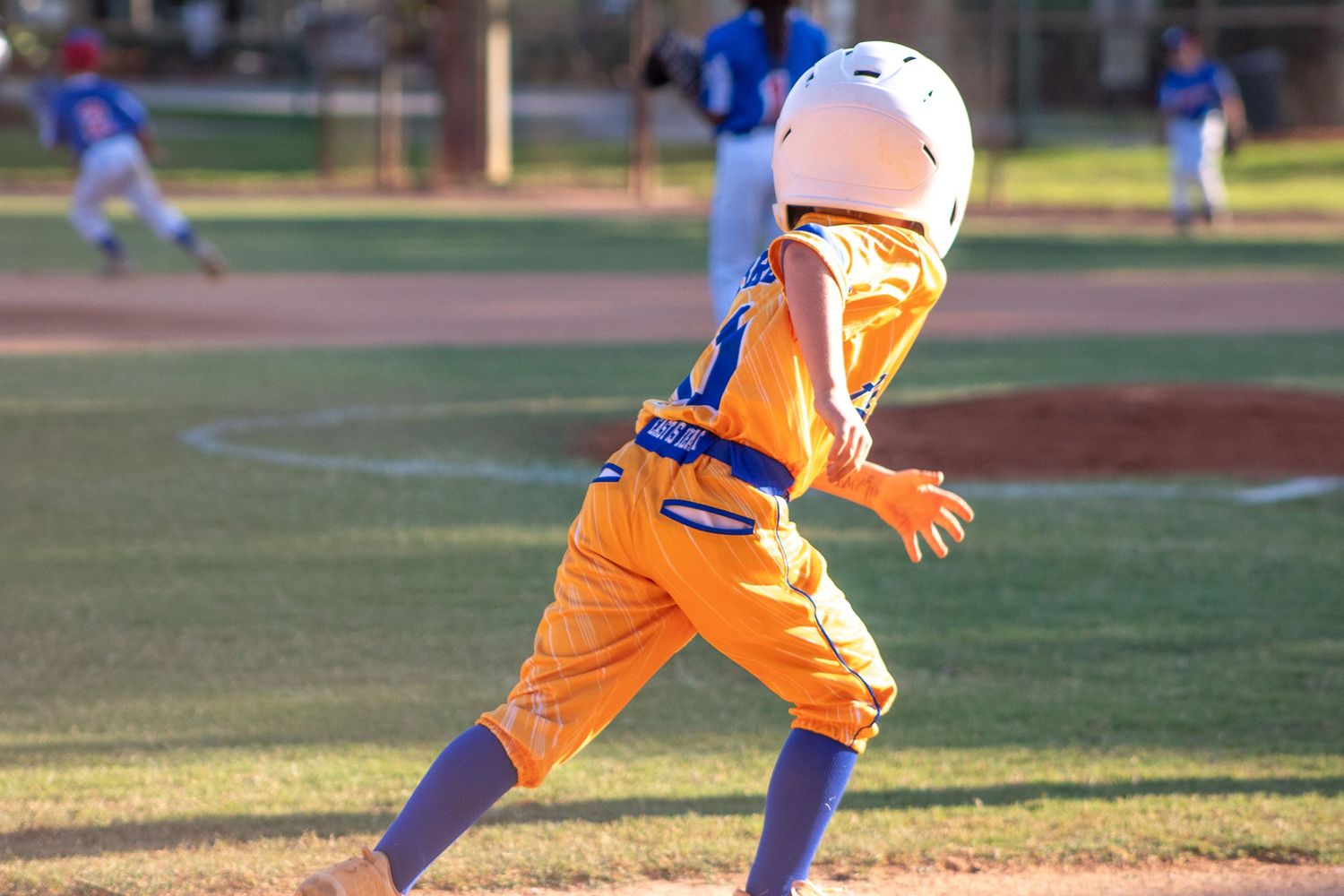 Youth baseball player in orange uniform runs on field during sunset game.