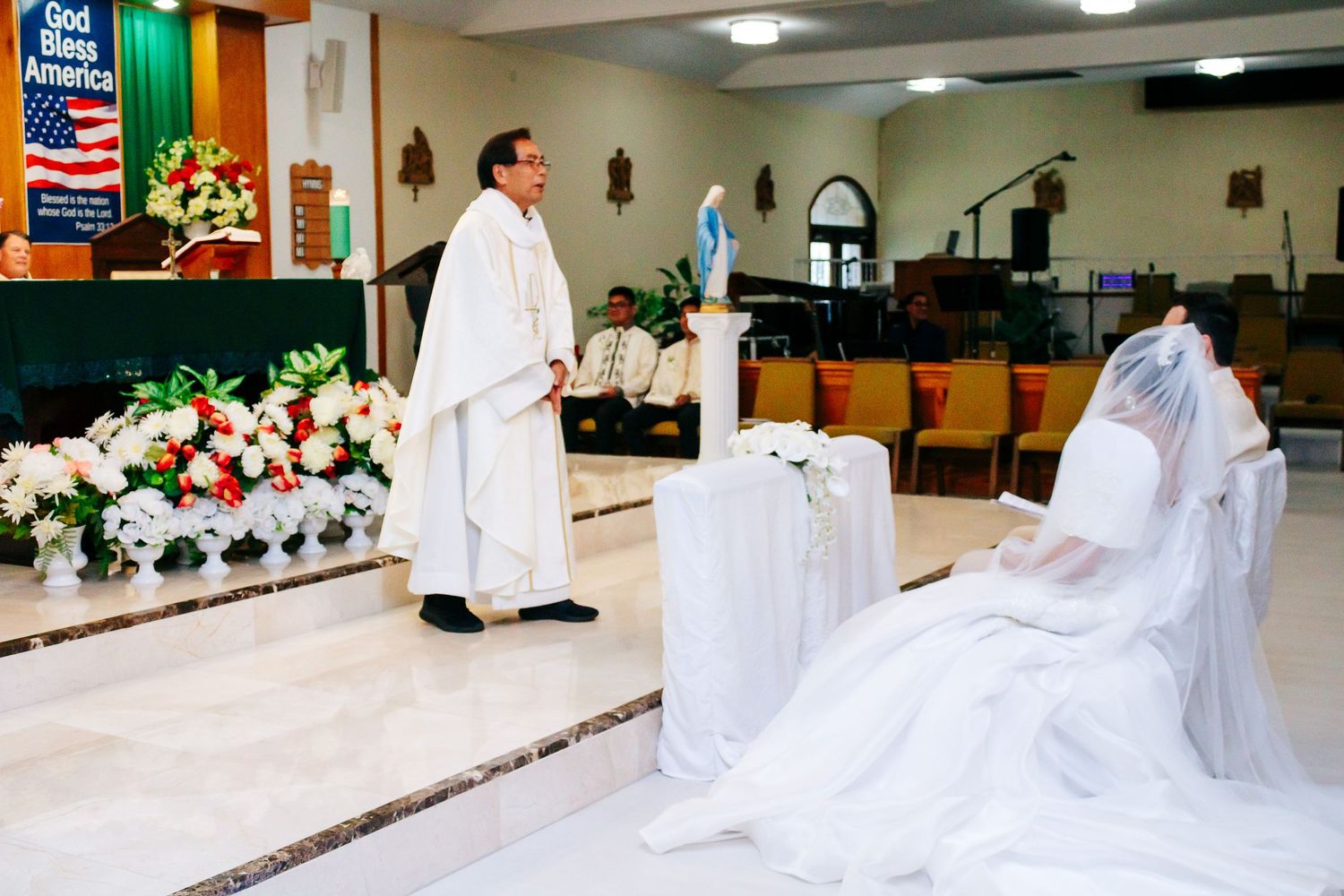 Priest in white robes conducts wedding ceremony at decorated church altar.