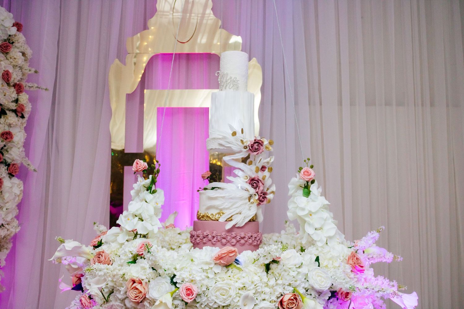 Multi-tiered white wedding cake with floral decorations against purple-lit backdrop.