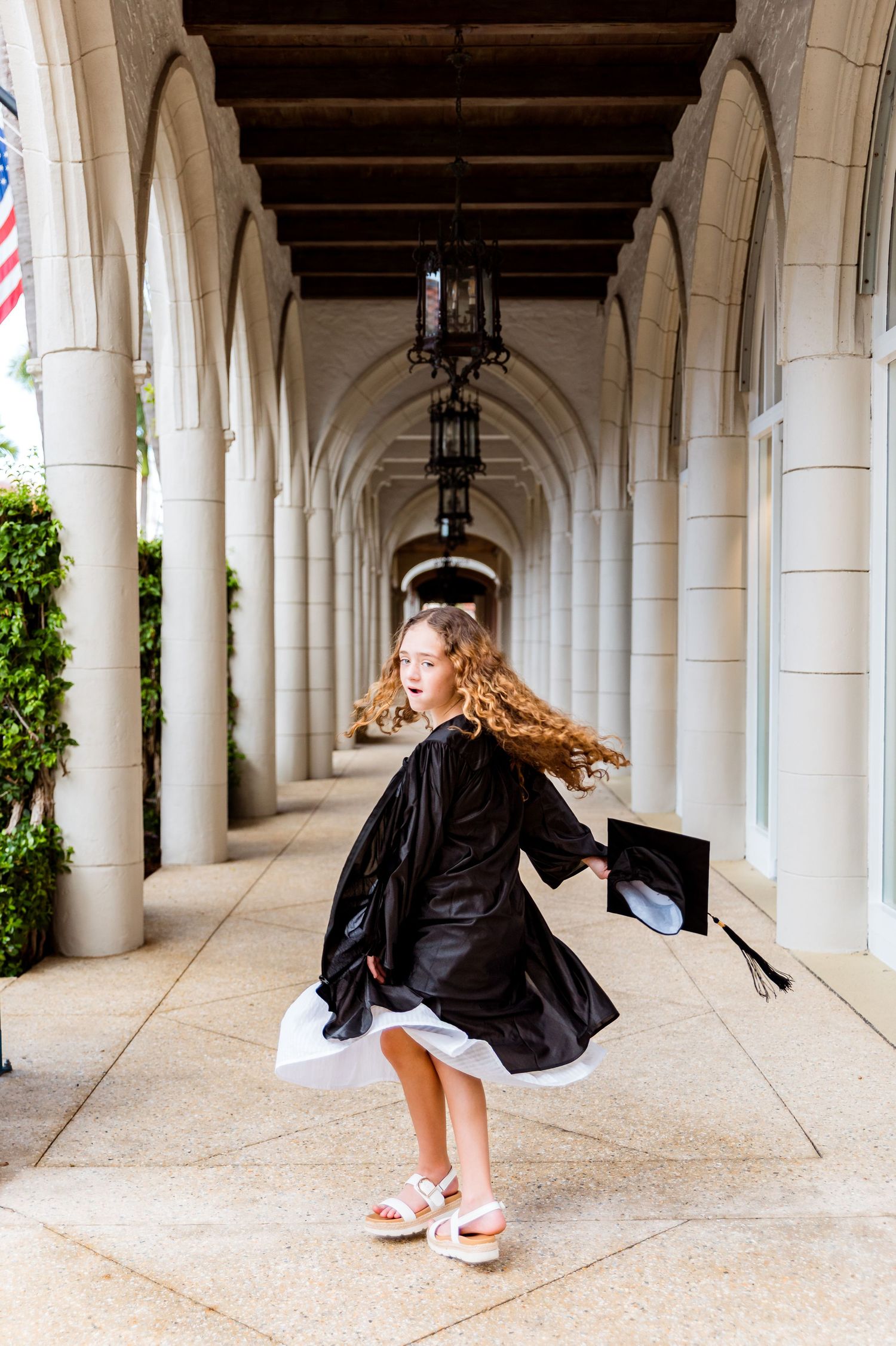 Young girl in black graduation gown twirling in an elegant arched colonnade, holding her mortarboard cap.