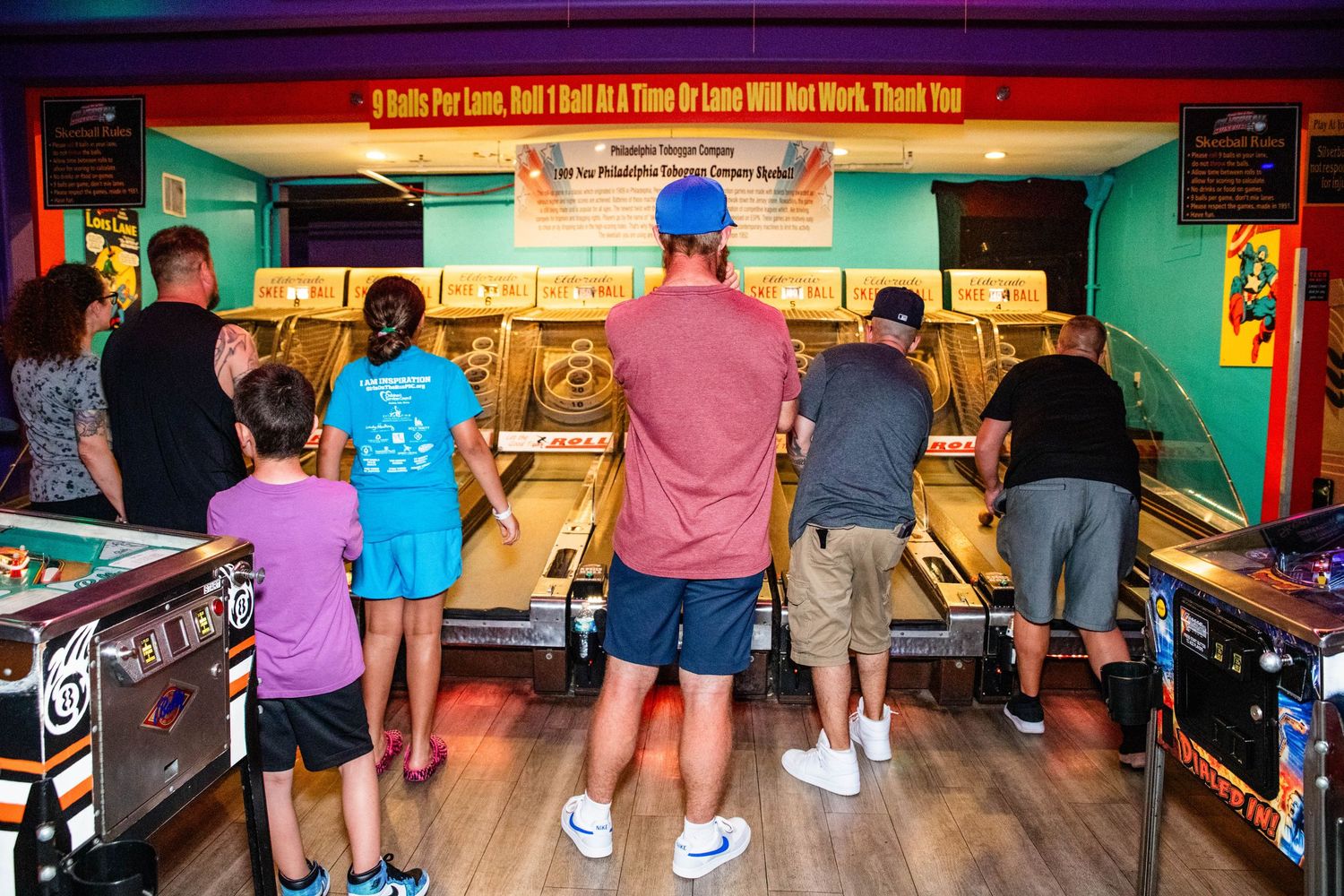 Several people are standing and playing various arcade games in a brightly lit and colorful arcade setting.