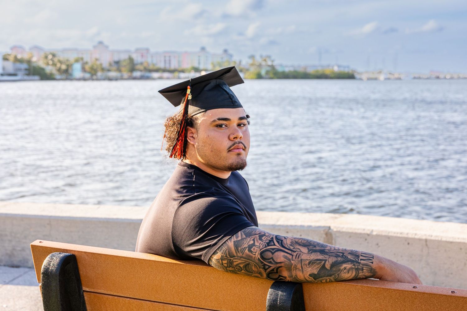 Young graduate wearing cap on bench near water, displaying tattoos, with coastal cityscape behind.