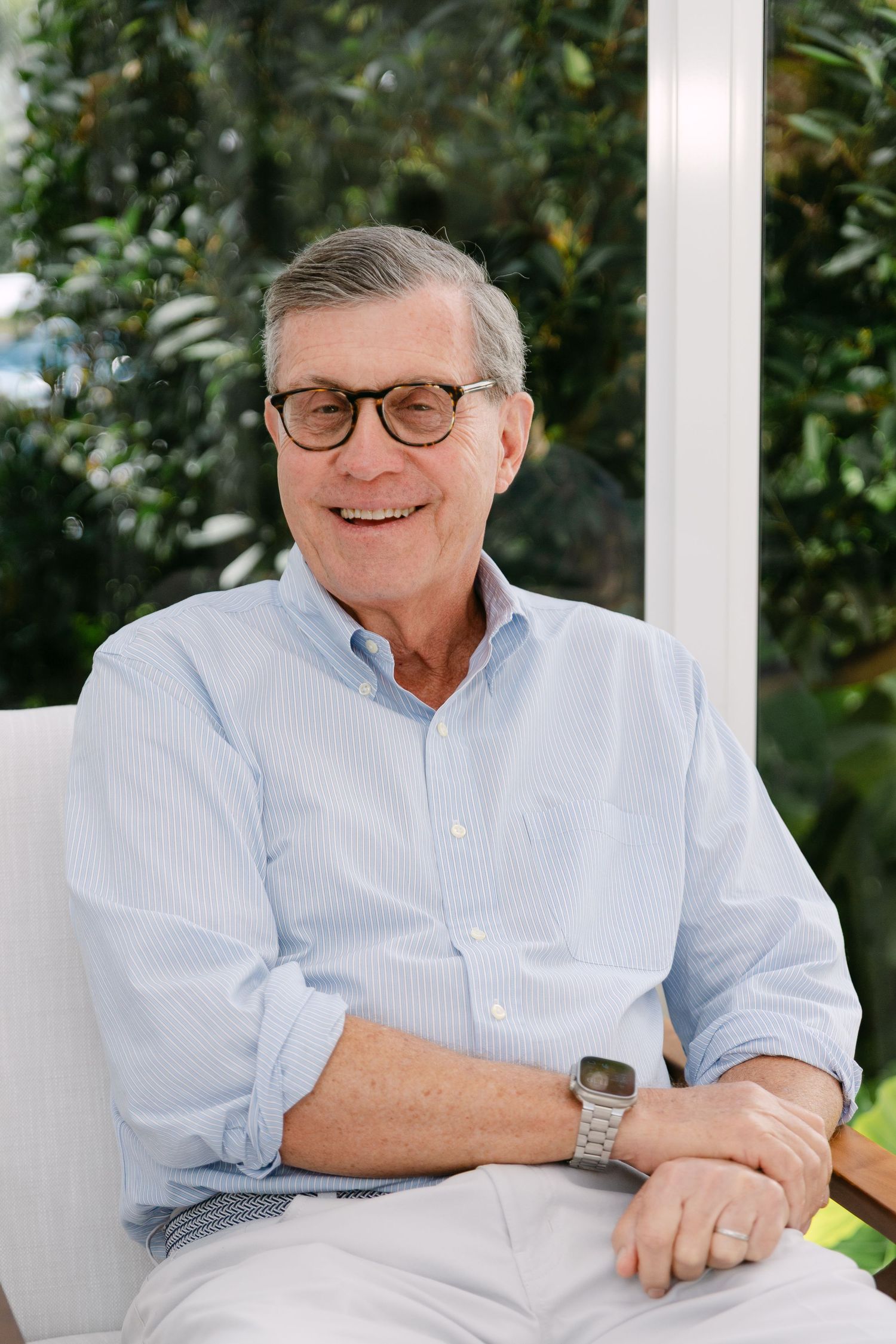 Smiling senior man with glasses in light blue shirt seated outdoors, surrounded by lush green foliage.