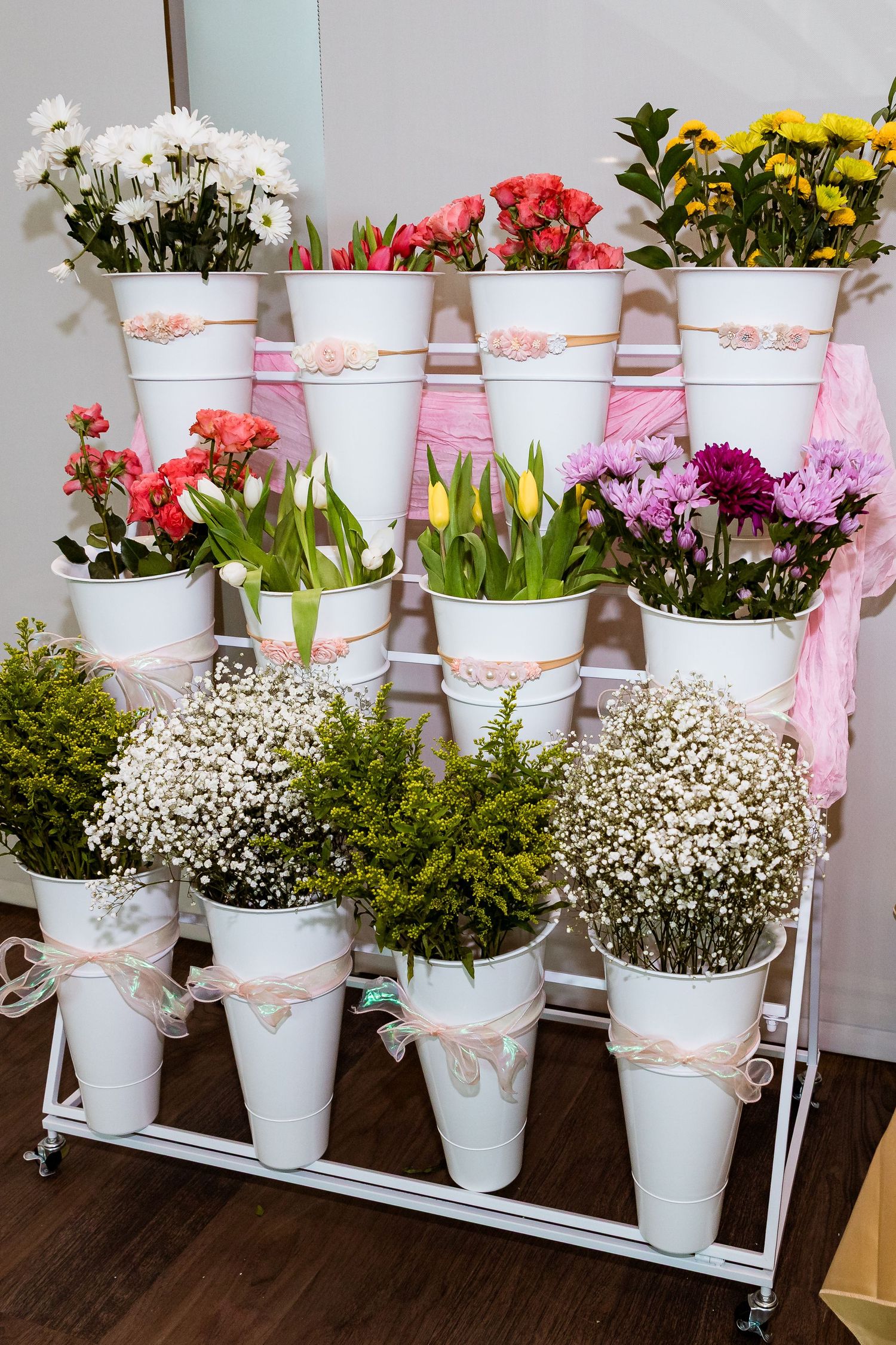 Tiered white metal stand displaying various colorful flowers in white decorative pots with ribbon accents.