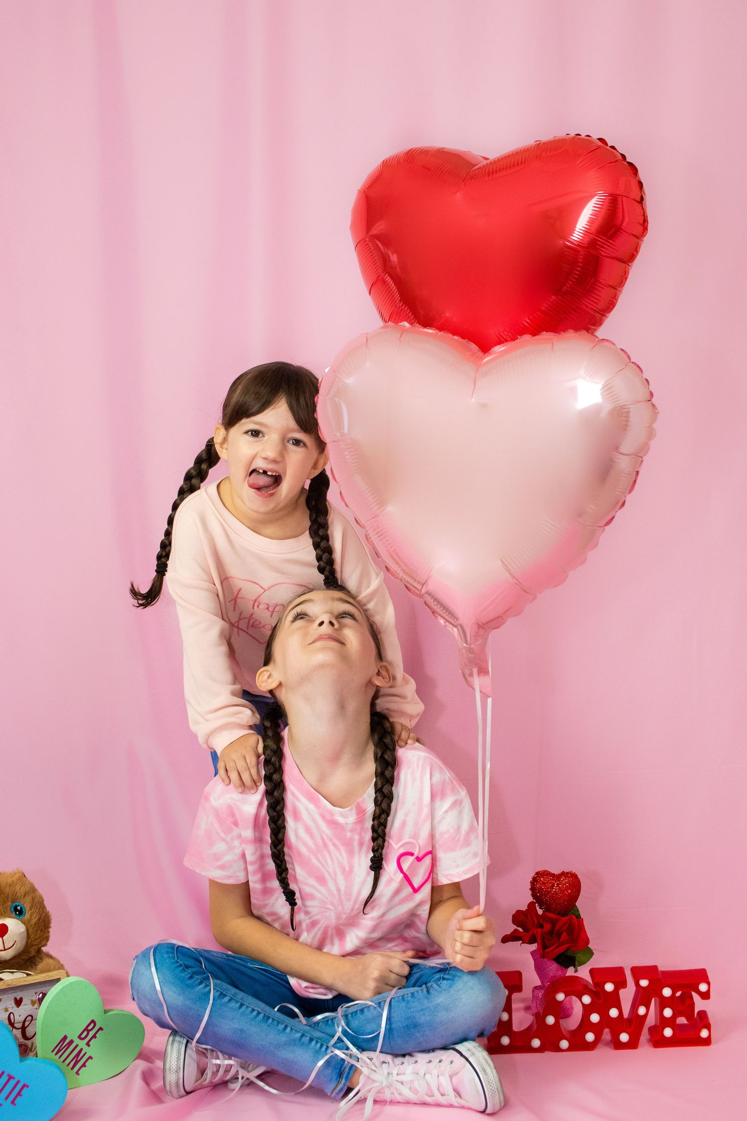 Two children playing and laughing with heart balloons and gifts on pink background.