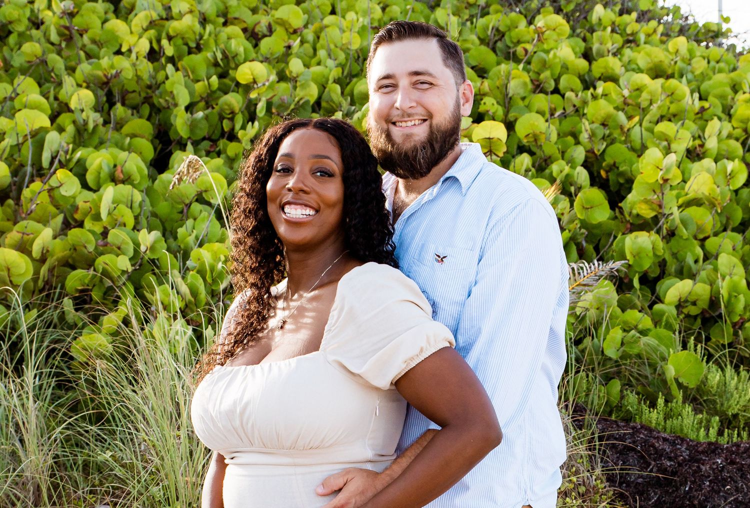 A couple embraces and smiles in front of lush green foliage on a sunny day.
