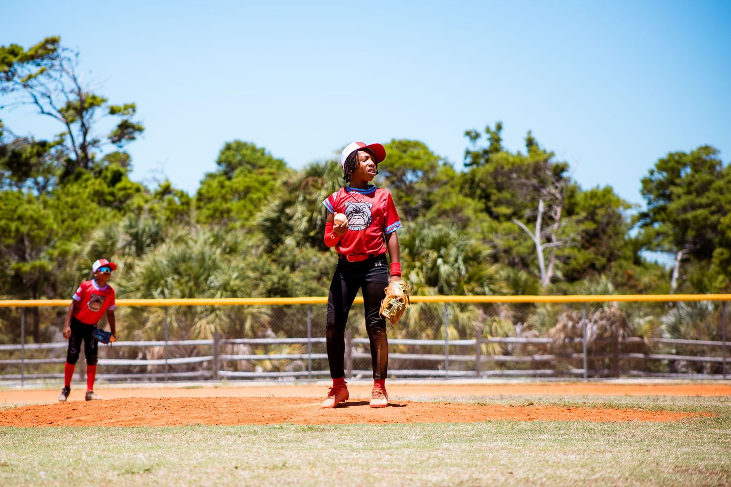 Pitcher in red uniform stands on mound against blue sky and trees background.