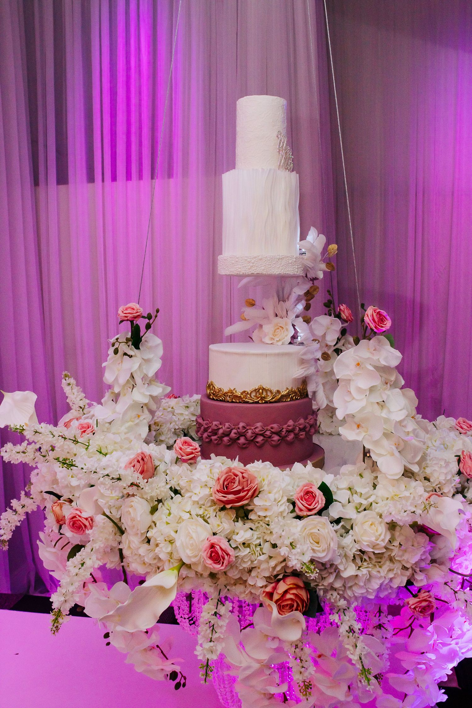 An elegant wedding cake display with cascading white orchids and pink roses under purple lighting.