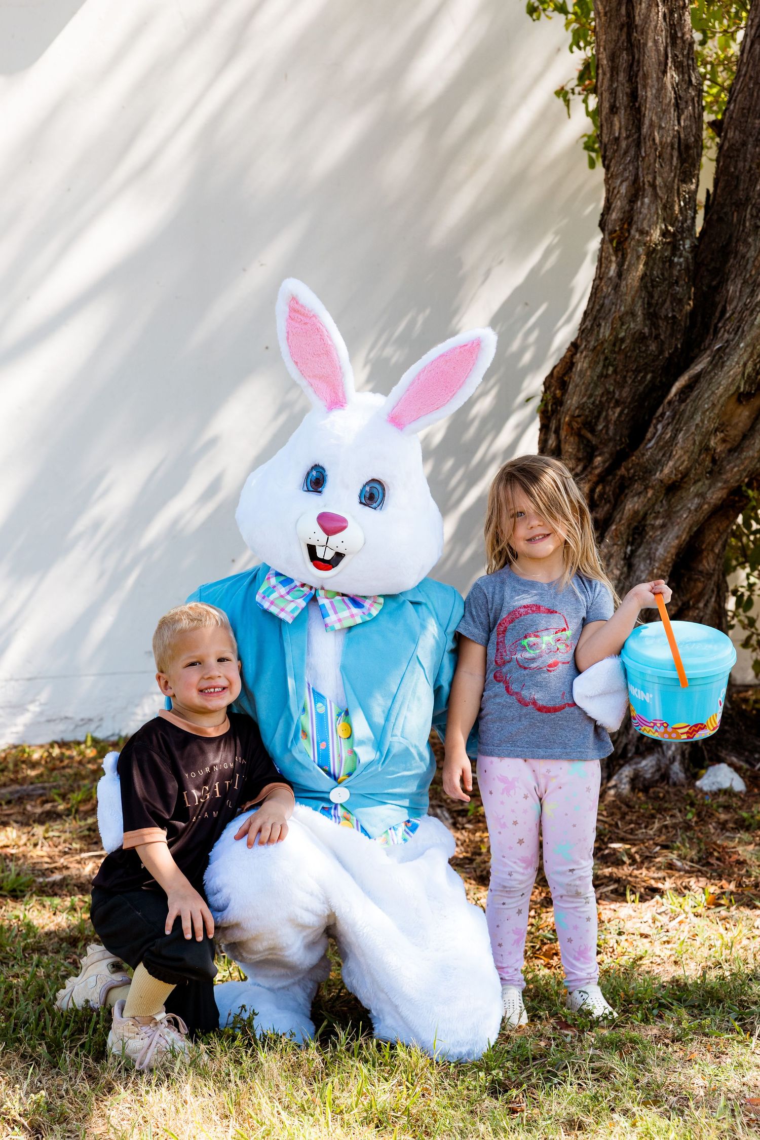 The Easter Bunny in a blue jacket poses with children holding Easter baskets outdoors.
