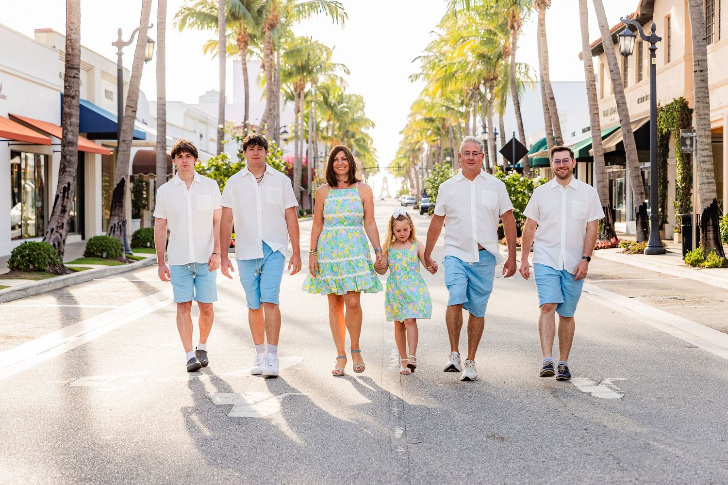 A group of people walking together down a palm tree-lined street in coordinated blue and mint green outfits on a sunny day.