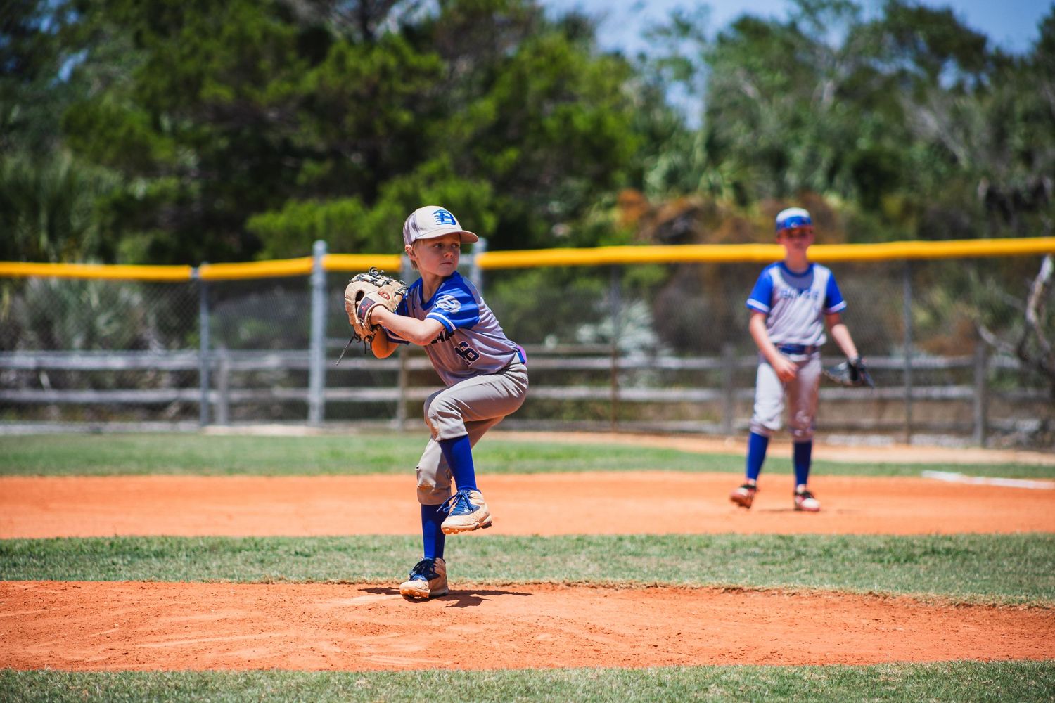Young pitcher in blue uniform winds up for a pitch on dirt mound during baseball game.
