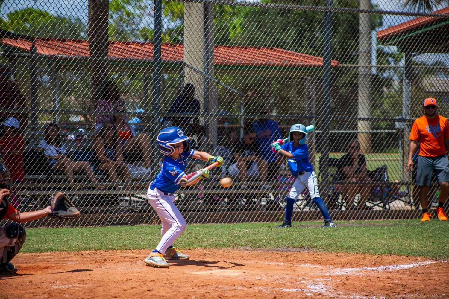 Baseball player at bat swings at pitch during little league game while teammates and spectators watch from dugout area.