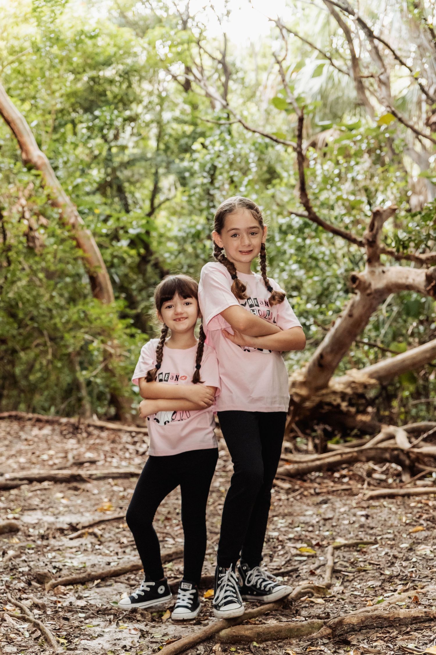 Two young children sit and play together on fallen tree branches in a sunlit wooded forest area.