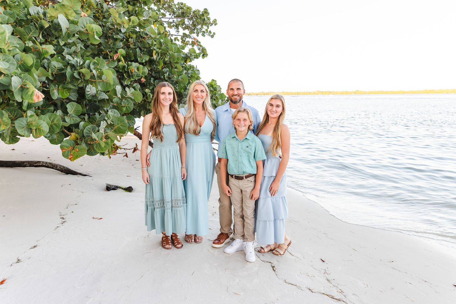 Beach family portrait session with group in aqua blue outfits standing by ocean at sunset.