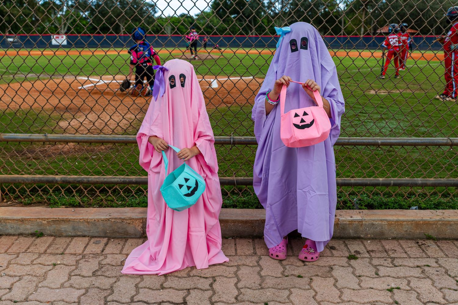 A group in pink and purple costumes stand together at a baseball field behind a chain link fence.