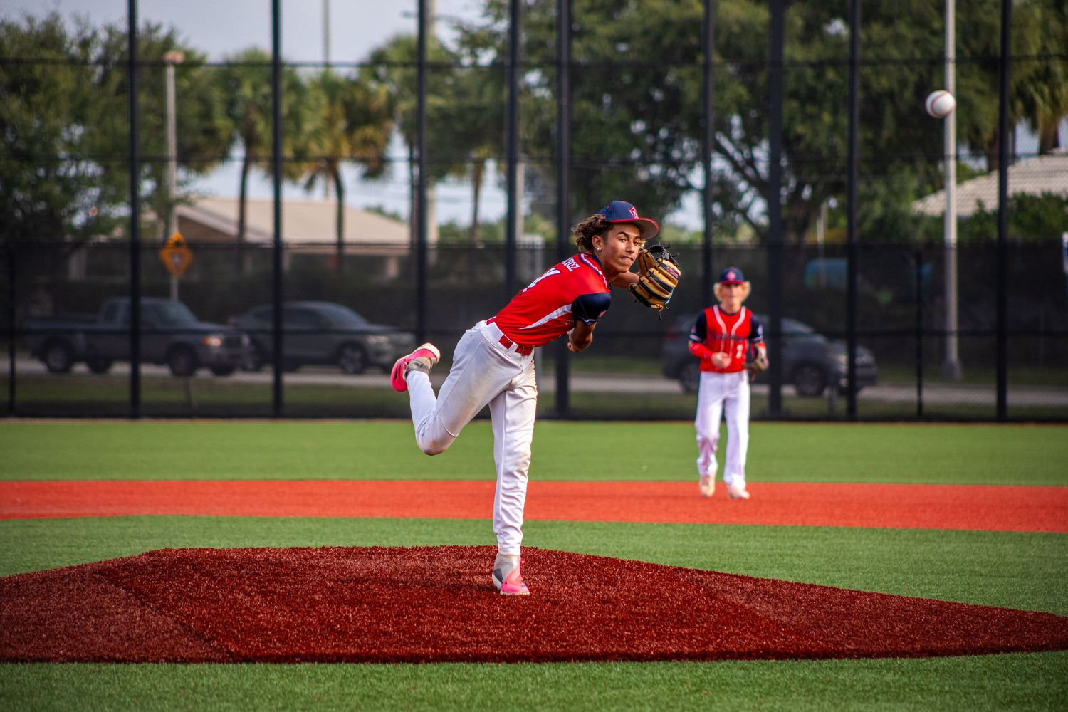 Young baseball pitcher in red uniform mid-throw on mound, with teammate visible in background on sunny field.