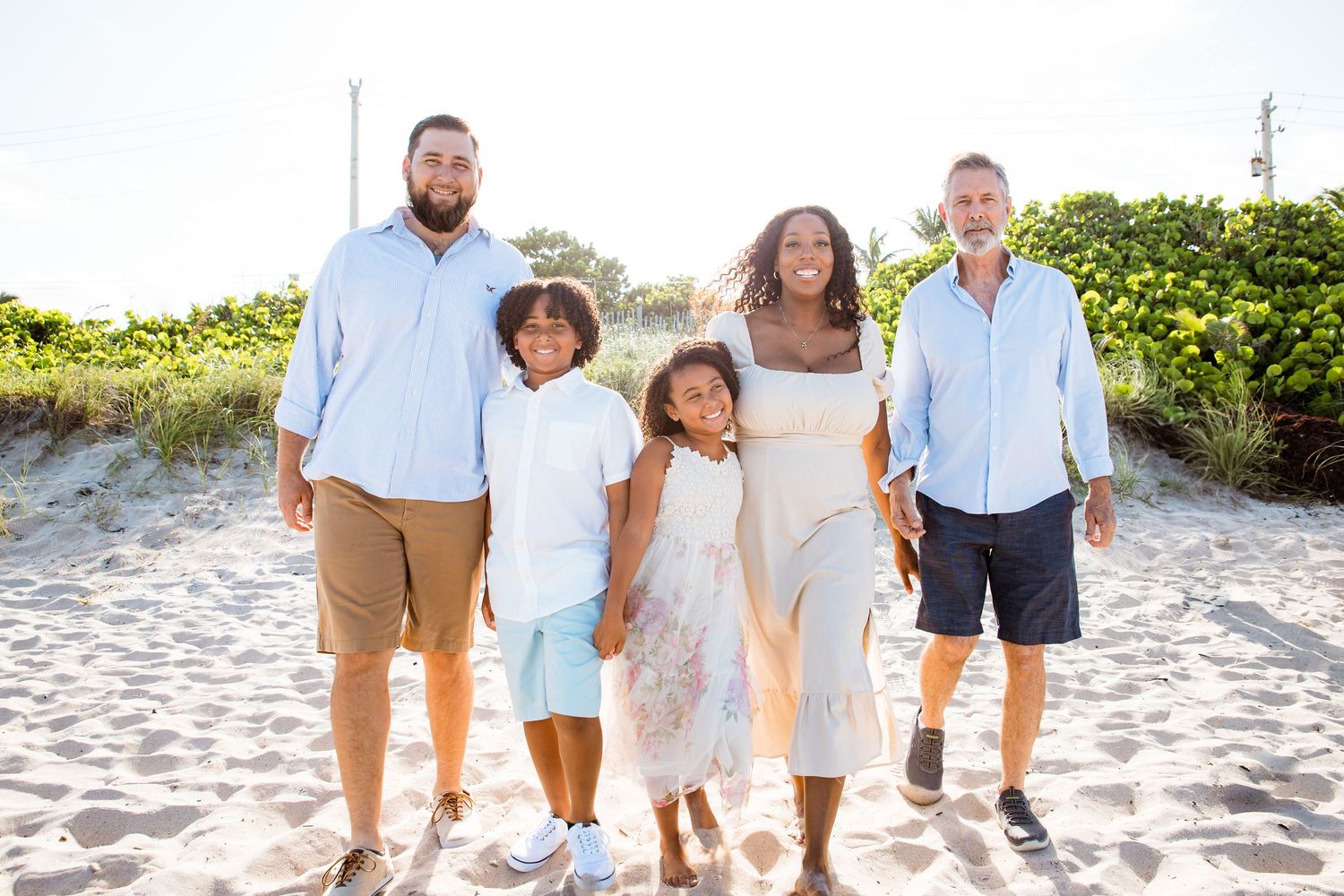 A family group wearing coordinated light-colored outfits walks together on a sunny beach with greenery in the background.