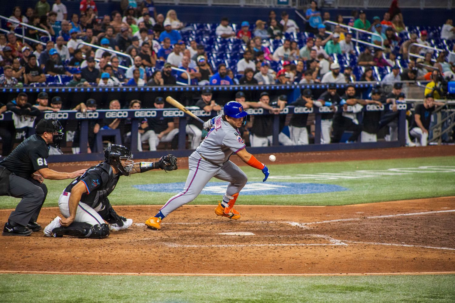 Baseball player swings bat during game with crowd visible in background at professional stadium.