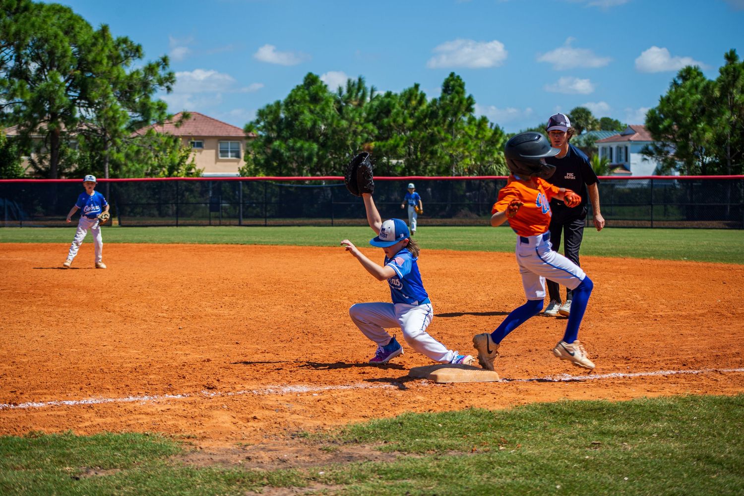Baseball players compete at home plate during a game on a sunny day with lush green trees in the background.