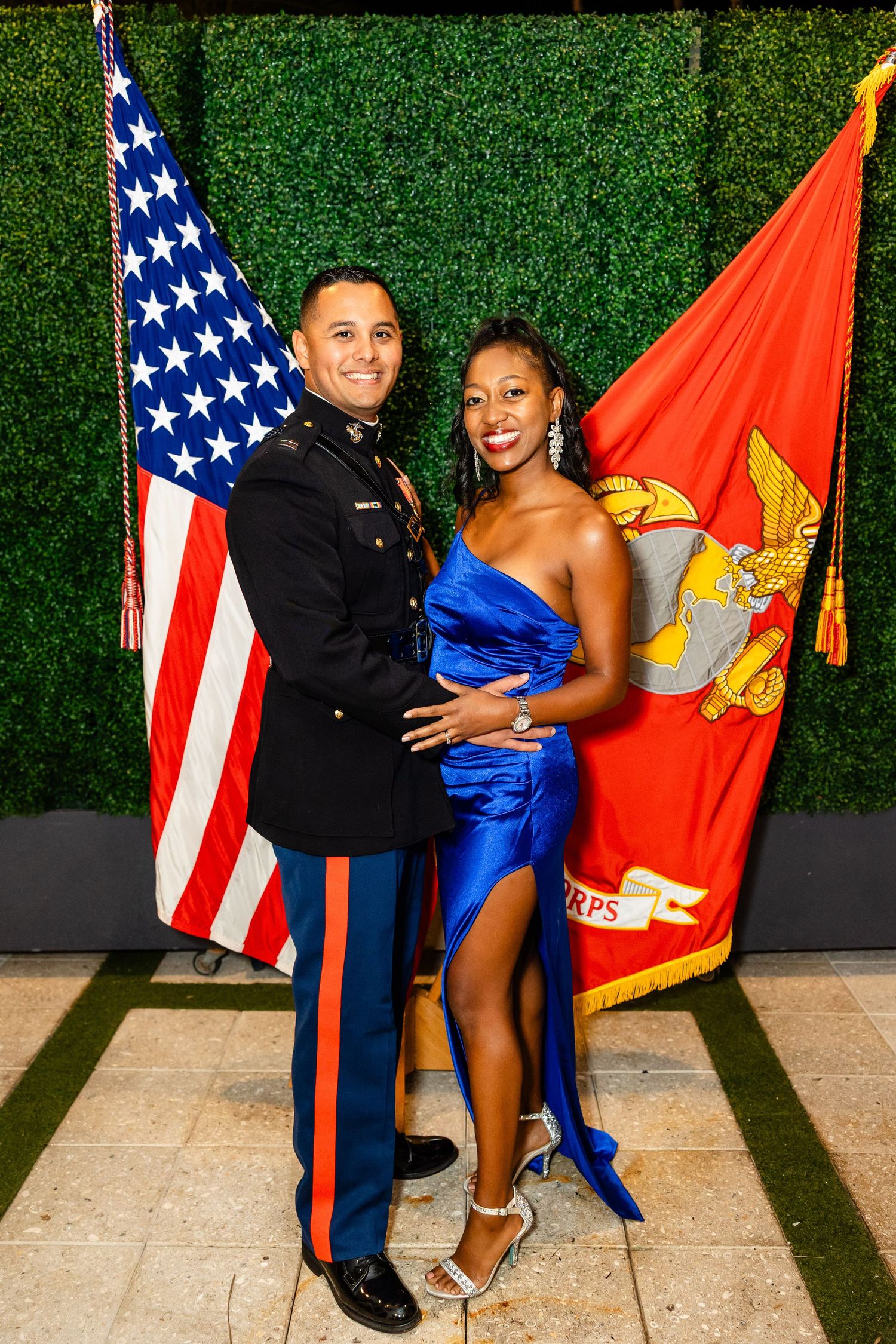 Marine in dress blues poses with partner in blue gown before US and Marine Corps flags.