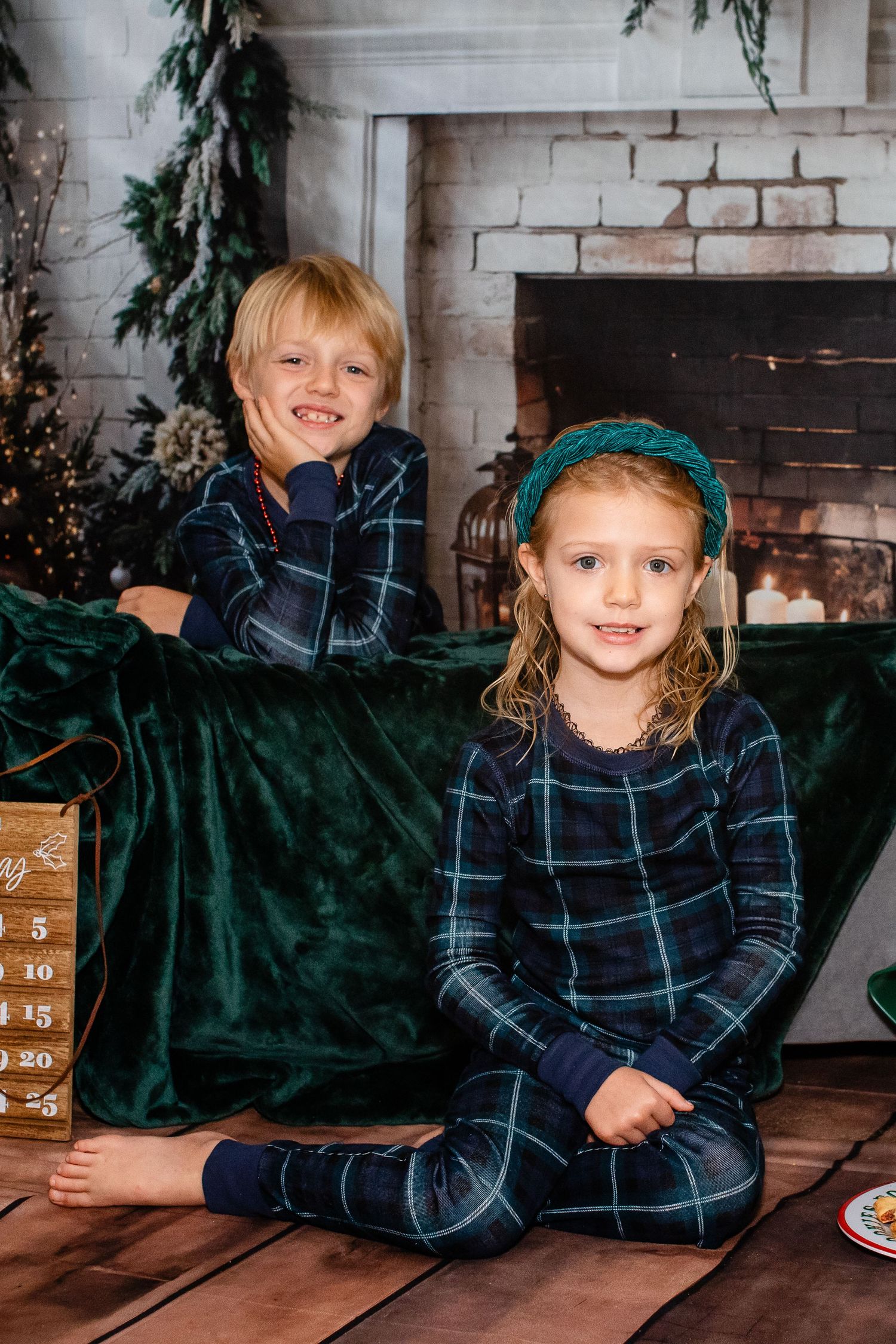 Two children in navy plaid pajamas pose in front of a decorated holiday fireplace setting.