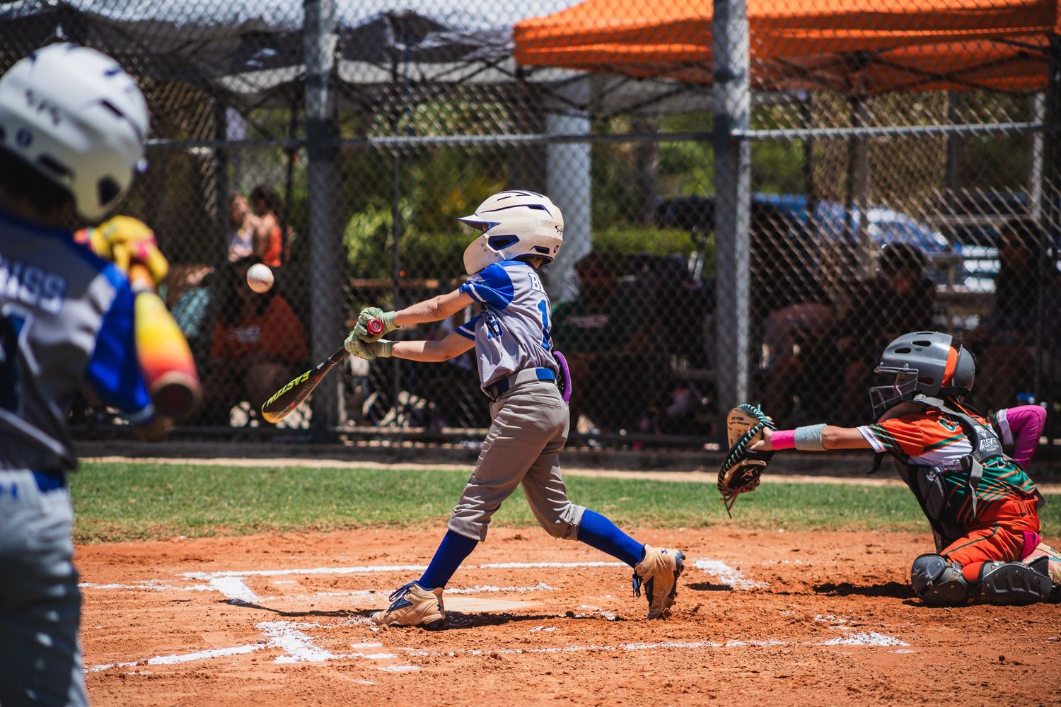 Youth baseball batter swings at pitch during game while catcher crouches behind home plate.