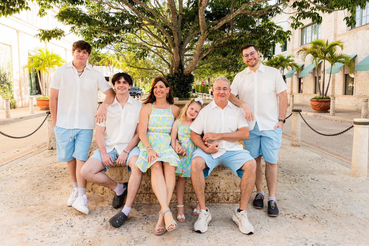 A multi-generational group wearing coordinated white and mint outfits sits together on a stone bench in a sunny outdoor setting.