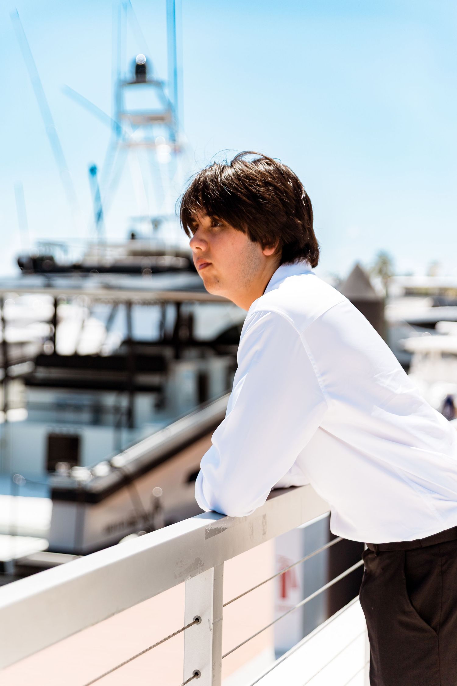 A person in white shirt leans on railing at marina with boats and blue sky in background.